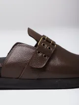 Mules de gamuza color tostado con diseño de tira ajustable con hebilla metálica y detalles de ojalillos decorativos. Posee plantilla ergonómica con logo grabado y suela plana de goma negra.