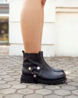 Bota de caña alta confeccionada en cuero negro, con suela track de goma. Presenta un diseño versátil con dos hebillas ajustables en la parte superior de la caña y una correa decorativa con tachas metálicas y argollas en la zona del tobillo.