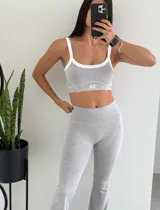 Top deportivo color gris melange, corte bralette con tirantes anchos blancos y ribete blanco en escote y sisas. Presenta costuras decorativas en blanco y un pequeño logo blanco en el centro inferior.