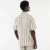 Camisa de manga corta color blanco con diseño de rayas verticales oscuras, cuello con muesca, cierre frontal con botones y bolsillo aplicado en el pecho.
