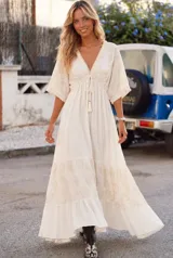 Vestido largo color blanco con bordados artesanales en tono beige, escote en V con cordones y borlas, mangas 3/4 estilo murciélago y diseño amplio con volados en la falda.