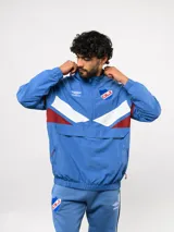 Buzo deportivo de nylon con medio cierre frontal y cuello alto. Presenta un diseño de bloques de color en azul, blanco y bordó, con el escudo del Club Nacional de Football y el logo de Umbro en el pecho.