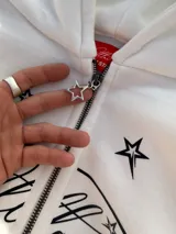 Campera canguro blanca con cierre frontal, capucha y bolsillos tipo kankaro. Presenta un estampado frontal con un óvalo con texto cursiva rodeado de estrellas negras.