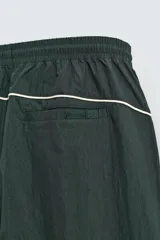 Pantalón jogger de corte relajado confeccionado en tejido técnico de color verde oscuro. Presenta cintura elástica con cordón interior, bolsillos laterales con cierre y bajo ajustable con elástico. Incluye detalles de líneas en contraste a lo largo de las piernas.