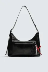 Bolso tipo bandolera de cuero sintético negro con acabado brillante. Presenta un compartimento principal con cierre de cremallera, un bolsillo frontal con cierre y detalle de charms colgantes decorativos. Incluye asa de hombro ajustable.