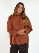Camisa de algodón color terracota con cuello alto tipo mao con volados, cierre frontal oculto y mangas largas con puños amplios y fruncidos.