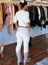 Pantalón de corte recto y tiro medio en color blanco, confeccionado en tejido elástico. Presenta cierre frontal con botón y cremallera, bolsillos laterales y viene acompañado de un cinturón fino de cuero sintético negro con hebilla metálica.