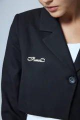 Saco sastrero corto a la cintura, color negro, con cierre frontal de un botón grabado, solapas clásicas, bolsillo ojal en el delantero, chapa metálica con logo en el pecho y bordado decorativo en la manga.