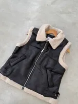 Chaleco de cuero sintético negro con interior y bordes de corderito sintético color crema. Presenta cierre frontal metálico, bolsillos laterales y correas ajustables con hebilla en el ruedo.