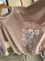 Buzo de algodón color tostado, de corte cropped y estilo oversize. Presenta cuello redondo, mangas largas con puños ajustados y elástico en la cintura. Destaca por una estampa exclusiva de flores en el frente y en una de las mangas.