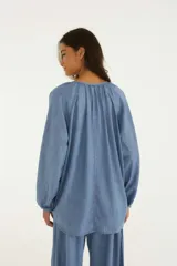 Blusa de mezclilla ligera con escote en V pronunciado, mangas largas amplias con puños ajustables mediante cordón y detalles de tiras colgantes en el cuello.