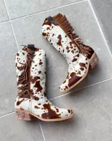 Botas texanas de caña media confeccionadas en cuero vacuno con estampado animal print tipo vaca. Presentan flecos laterales en gamuza marrón, taco cuadrado bajo y puntera ligeramente redondeada.