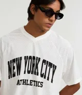 Remera de hombre con corte boxy, confeccionada en tejido mesh (malla) blanco, con cuello en V y estampa arqueada en negro que dice "NEW YORK CITY ATHLETICS".