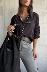 Camisa de color negro con cuello clásico y cierre frontal de botones blancos. Presenta mangas largas con detalles bordados en los puños.