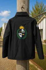 Campera impermeable de tela taslon color negro, con diseño de cuello alto y cierre frontal. Presenta un gran bordado en la espalda que incluye el escudo nacional uruguayo y el logo de la marca Bagual, además de un parche circular con la bandera de Uruguay en la manga.