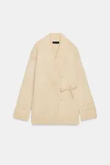 Chaqueta cruzada de punto color beige con manga larga, cuello smoking y cierre frontal con lazada. Presenta detalles de calados en los puños.