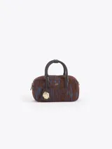 Cartera tipo baulito de cuero descarne vacuno con textura rayada en tonos marrón y azul. Presenta manijas cortas, cierre superior y un charm metálico dorado en forma de mariquita colgando de un tag de cuero. Incluye correa larga desmontable y regulable.