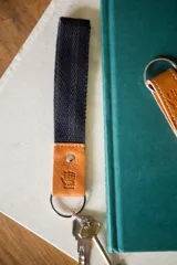 Llavero con correa de algodón color negro y aplique de cuero color marrón con logo grabado. Incluye argolla metálica para llaves.