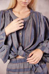 Camisa de lino con cuello mao y cierre frontal de botones. Presenta mangas largas con frunces que aportan volumen y un diseño de rayas verticales en tonos azul, gris y naranja.