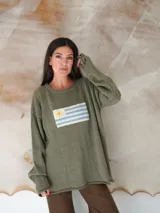 Sweater de hilo de algodón en color beige con diseño tejido de la bandera de Uruguay en el centro. Presenta cuello redondo, mangas largas y un corte holgado.