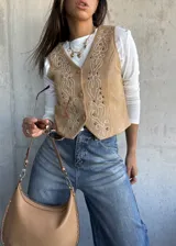 Chaleco de gamuza color beige con bordados decorativos en hilo blanco y apliques de tachas metálicas. Presenta cierre frontal con botones y diseño de corte clásico.