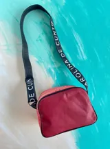 Bandolera Kaira de la línea Ottenta de Carolina De Cunto, confeccionada en cuero color rojo con diseño estilo splash en negro y bolsillo frontal superior en tono cobre. Cuenta con correa larga regulable negra con el nombre de la marca en blanco y cierre superior con cremallera.