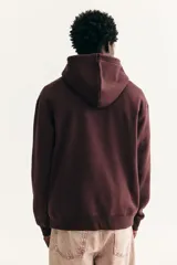 Canguro de corte holgado en color bordó, con capucha ajustable mediante cordones, hombros caídos y bolsillo frontal tipo canguro. Presenta un pequeño estampado central de un jarrón con flores coloridas.