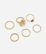 Kit de seis anillos dorados con diferentes diseños y acabados, incluyendo un anillo con piedra marrón, uno con detalle de lazo, uno con textura trenzada y anillos lisos y con cuentas.