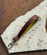 Llavero de cuero en formato de tira con diseño minimalista, herrajes metálicos color bronce viejo y gancho tipo mosquetón. Confeccionado en cuero genuino con costuras pespunteadas.
