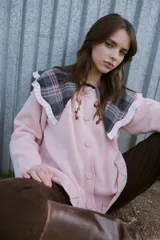 Cardigan de punto color rosa con cierre frontal de botones tejidos. Presenta un cuello grande estilo babero con estampado de cuadros escoceses en tonos gris y rosa, con terminación de volados. Incluye detalles bordados de flores en la parte superior delantera y calados decorativos en el ruedo y los puños.