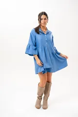 Vestido corto azul, estilo camisero, con cuello camisero, cierre frontal con botones y mangas acampanadas con volados. Presenta un diseño holgado con cortes fruncidos en el cuerpo.