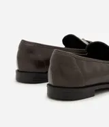 Mocasín Satinato de poliuretano color marrón oscuro, con punta redondeada, tacón bajo y empeine decorado con tiras cruzadas, hebilla ajustable y ojetes metálicos.