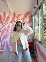 Campera de algodón premium color crudo con detalles en marrón y rosa. Presenta un cierre frontal, cuello redondo, mangas largas acampanadas y un logo bordado en el pecho.