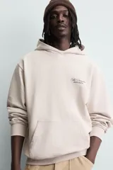 Sudadera hoodie relaxed fit de color beige, con cuello con capucha, manga larga y bolsillo frontal tipo canguro. Presenta un pequeño estampado de texto en contraste en el pecho.
