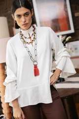 Blusa blanca de textura arrugada, con cuello alto, abertura frontal tipo gota y mangas largas acampanadas con detalle de frunces.