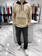 Canguro oversize beige con capucha y cordones negros, cierre frontal parcial con cremallera, bolsillo canguro delantero con solapa y pequeño bordado negro en el pecho.