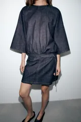 Vestido corto de denim azul oscuro, con cuello redondo, mangas amplias de largo tres cuartos y cintura elástica con cinturón integrado del mismo tejido. Presenta costuras a contraste y cierre posterior con cremallera oculta.
