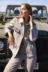 Chaqueta de denim liviana color blanco con volados en el frente, cuello camisero y cierre frontal de botones. Presenta un calce relajado y bolsillos funcionales.