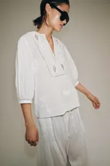 Camisa blanca de algodón con escote en pico, mangas por debajo del codo y detalles de bordados y encaje a tono en el pecho y las mangas.