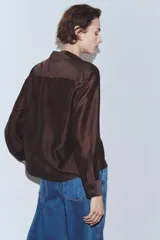 Camisa de manga larga color marrón oscuro, con cuello solapa, escote pico y bajo asimétrico. Presenta un bolsillo plastrón en el delantero con bordado combinado a tono.