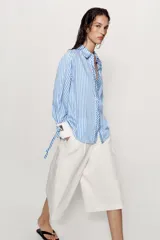 Camisa de cuello solapa y manga larga con rayas verticales celestes y blancas. Cuenta con puños anchos blancos contrastantes, ajustables con hebilla dorada y lazos, y cierre frontal con botones dorados.