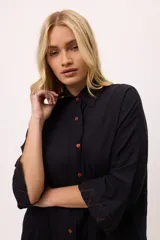 Vestido camisero negro de algodón, con mangas tres cuartos y cuello camisero. Presenta detalles de costura en zigzag color naranja en el cuello y en los puños.