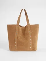 Cartera tipo shopper de gran tamaño confeccionada en material efecto gamuza color rojo. Presenta correas reforzadas decoradas con tachas metálicas plateadas y un dije colgante en forma de corazón con detalles de tachas. Cuenta con una placa metálica pequeña con logo en la parte inferior frontal.
