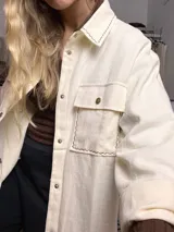 Sobre camisa color manteca con costuras decorativas en zigzag de color oscuro. Presenta cuello clásico, cierre frontal con botones metálicos a presión, mangas largas con puños abotonados y un bolsillo frontal con solapa.