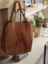 Bolso tipo tote de cuero color marrón, con diseño artesanal que presenta detalles de costuras trenzadas en los laterales y en el centro. Cuenta con dos asas superiores trenzadas y un tamaño amplio.