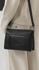 Bandolera de tamaño mediano en color gris, confeccionada en símil cuero con textura granulada. Presenta un bolsillo frontal con cierre, herrajes metálicos oscuros y correa ajustable y desmontable.