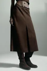 Falda midi de tejido efecto ante en color marrón oscuro, con tiro medio, trabillas en la cintura y cinturón ajustable con hebilla metálica. Presenta un diseño de bajo recto con una abertura frontal central y cierre lateral con cremallera oculta.