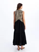 Vestido largo sin mangas, de color negro con la parte superior bordada en tonos dorados y negros con patrones geométricos. Presenta escote en V y falda con volados escalonados.