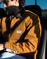 Campera de estilo workwear color camel, confeccionada en canvas resistente con interior de corderito sherpa. Presenta cuello de pana en color marrón oscuro, cierre frontal con botones metálicos color cobre, cuatro bolsillos frontales y un bolsillo interior.