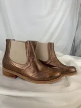 Botín tipo Chelsea de cuero metalizado en tono bronce, con paneles elásticos laterales en color beige. Presenta detalles de costuras decorativas tipo brogue en la puntera y laterales, con suela de goma antideslizante y taco bajo foliado.